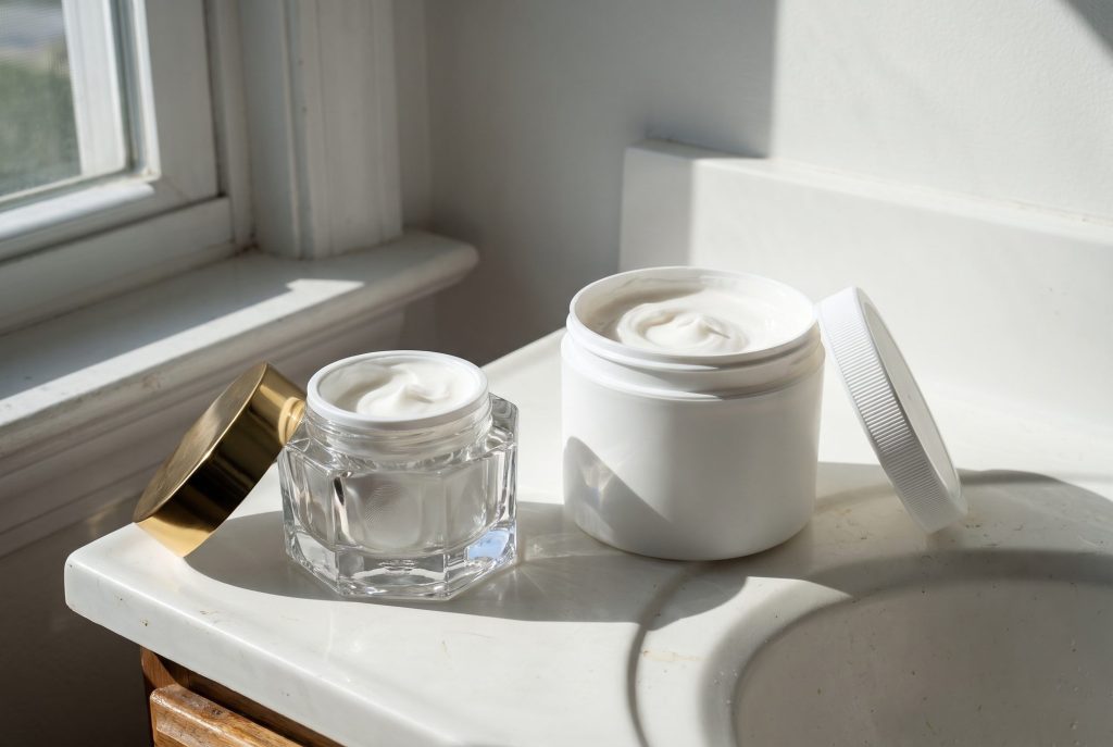 A small cut-glass jar with a gold lid next to a large plain plastic tub, both holding the same white cream