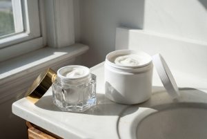 A small cut-glass jar with a gold lid next to a large plain plastic tub, both holding the same white cream