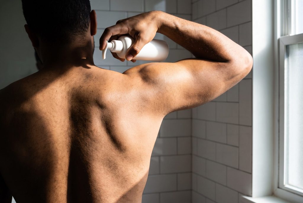 A man reaches over his shoulder with a pump bottle, trying to get to the middle of his back