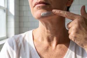 A fingertip spreads sunscreen on a woman's chin while her neck, with fine lines, stays bare