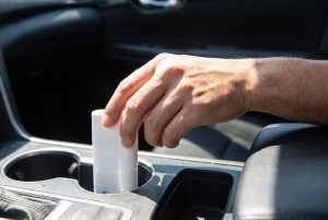 A hand reaches for a plain white sunscreen stick standing in a car's cup holder