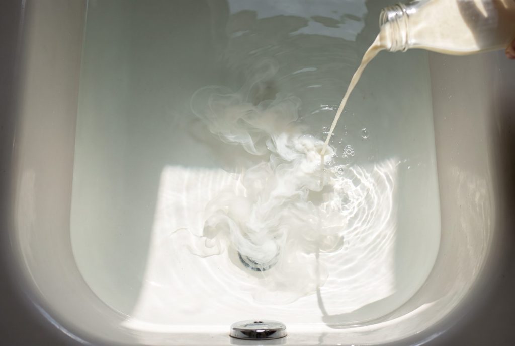 A thin stream of milk pours into a bathtub of clear water, unfurling into silky white wisps