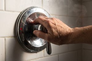 A wet hand turns a chrome shower valve toward the red hot mark as steam rises