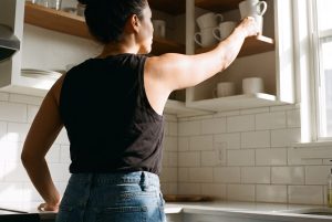A woman in a sleeveless black top reaches up to a high kitchen shelf of white mugs
